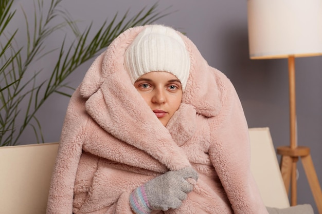 Closeup portrait of cold frozen woman wearing warm clothes and wrapped in coat sitting in room near lamp looking at camera needs to get warm feels icily in her house