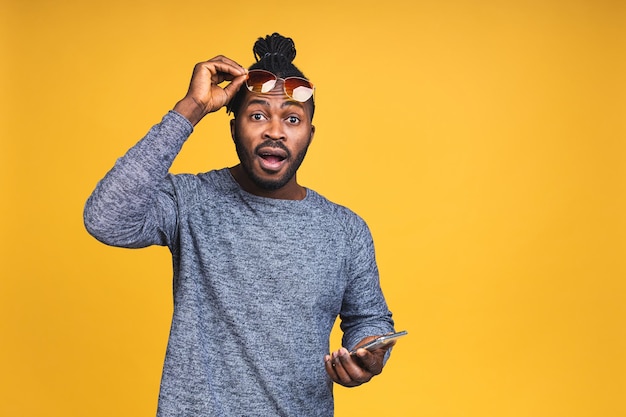 Closeup portrait of handsome young african american black man, shocked, surprised, wide open mouth, mad by what he sees on his cell phone, isolated on yellow background.