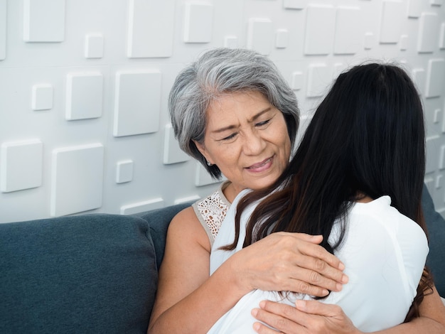 Closeup portrait of happy Asian senior mother or grandparent white hair embracing her beautiful daughter or grandchild with love care and comfort while sit on grey couch in living room at home