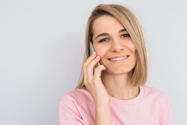 Closeup portrait of happy beautiful blonde female model glad to speak with friend via mobile phone posing over white wall Pretty Caucasian woman with pleased expression talks on smartphone People