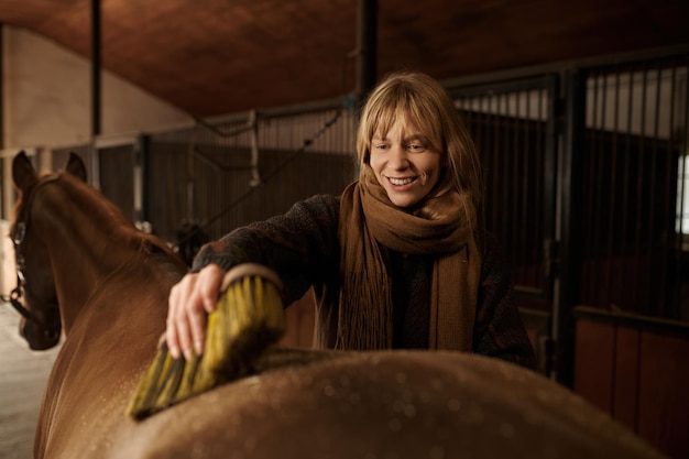 Closeup portrait of happy horsewoman cleaning her stallion with brush. Female rider washing horse before or after riding