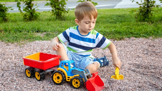 Closeup portrait of happy smiling 3 years old child boy digging sand on the playground with toy plastic truck or excavator