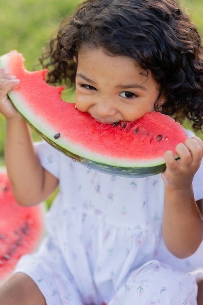 Closeup portrait of a little swarthy girl with curly hair eating a watermelon on the lawn