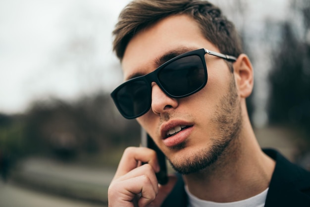 Closeup portrait of modern young man wearing black suit and sunglasses texting on the phone messages for his girlfirend in the street Technology and communication concept