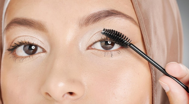 Closeup portrait of muslim woman applying mascara makeup to eyelashes Headshot of stunning arab model doing her beauty routine against grey background Face of beautiful woman extending her lashes