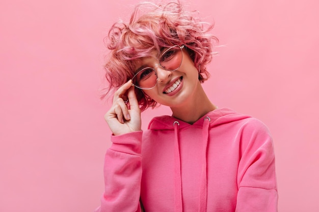 Closeup portrait of pinkhaired curly woman wearing sunglasses and looking into camera on isolated Charming curly girl in bright hoodie smiles on pink background