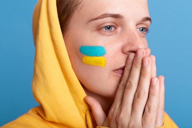 Closeup portrait of praying woman wearing yellow hoodie and with flag of Ukraine on her cheek standing with palms together looking away prays for Ukraine isolated over blue background