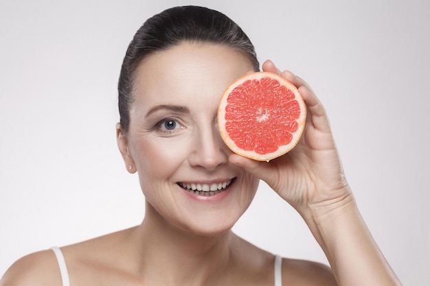 Closeup portrait of pretty woman with perfect skin smiling after cream, balm, mask, lotion, holding half of grapefruit in front of her eye, isolated on grey background. Indoor, studio shot,copy space