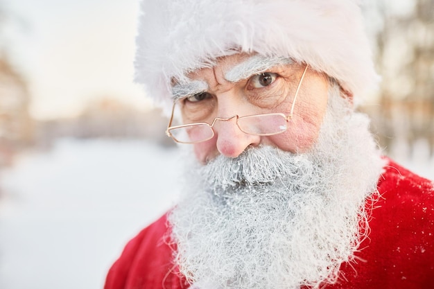 Closeup portrait of serious santa claus looking at camera over eyeglasses in winter setting outdoors