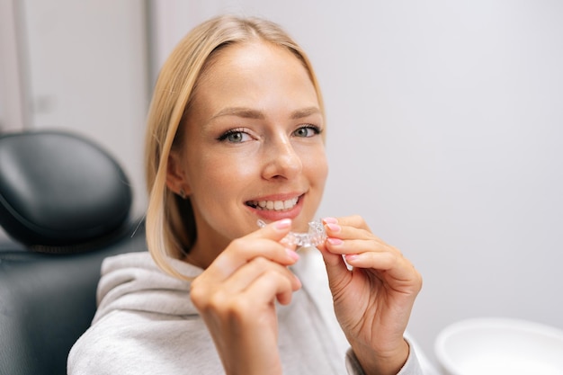 Closeup portrait of smiling blonde female patient holding invisible braces aligner sitting on chair in dentistry clinic Close up of mobile orthodontic appliance for dental correction