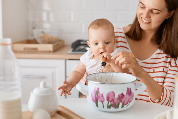 Closeup portrait of smiling positive female wearing striped shirt baking together with her toddler daughter sitting at table with pot and whist and mixing ingredients for dough