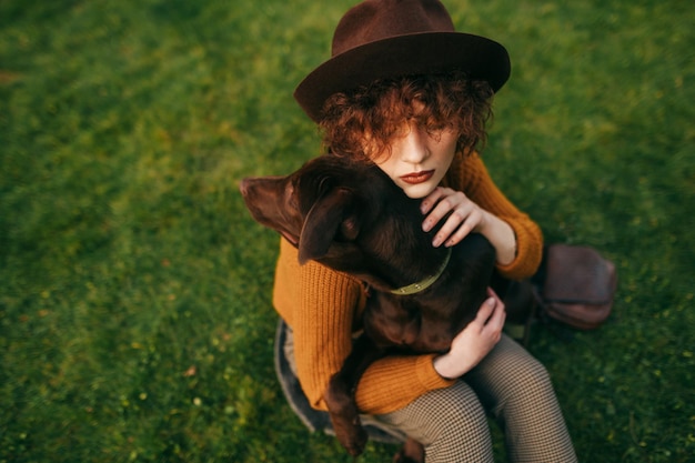 Closeup portrait of stylish lady in hat and with curly hair hugging dog on lawn background top view