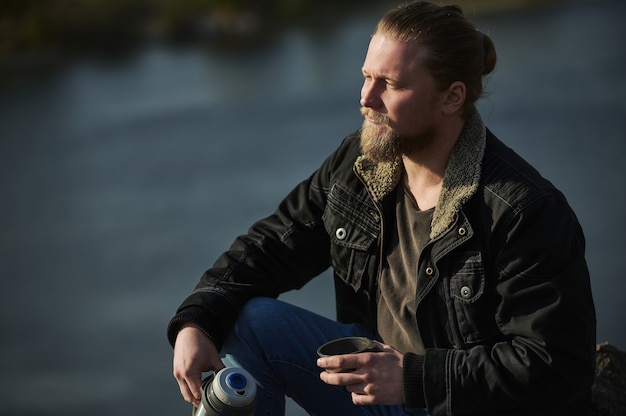 Closeup portrait of stylish lamber man holding thermos with hot drink , looking into distance and enjoying nature