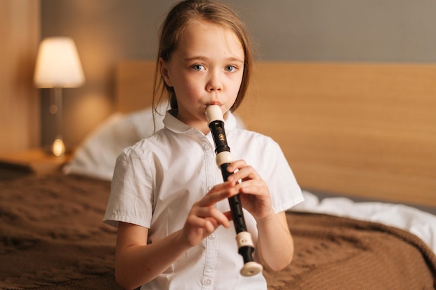 Closeup portrait of talented adorable little girl playing flute sitting on bed in bedroom looking at camera