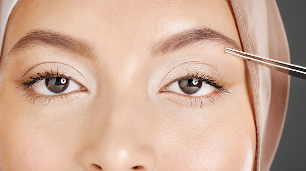 Closeup portrait of traditional muslim woman using tweezers to pluck her eyebrows Half headshot of confident arab model wearing a hijab in a studio Cultural middle eastern grooming her facial hair