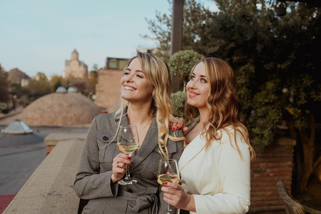 Closeup portrait of two female friends in strict suits laughing drinking wine on the terrace outside