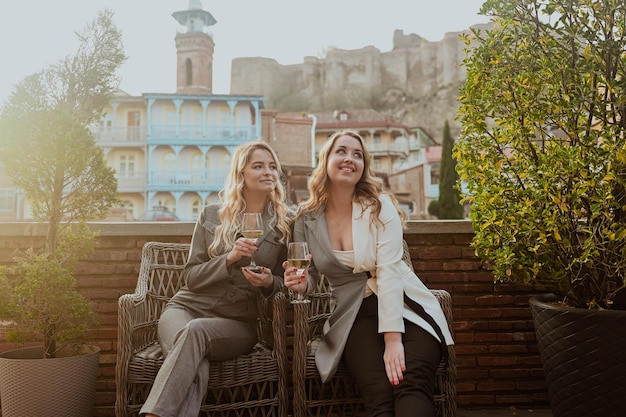 Closeup portrait of two female friends in strict suits laughing drinking wine on the terrace outside