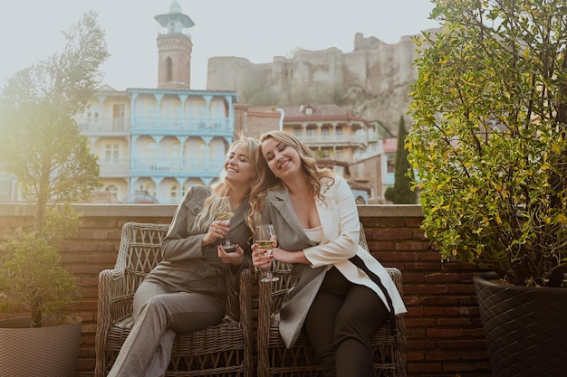 Closeup portrait of two female friends in strict suits laughing drinking wine on the terrace outside