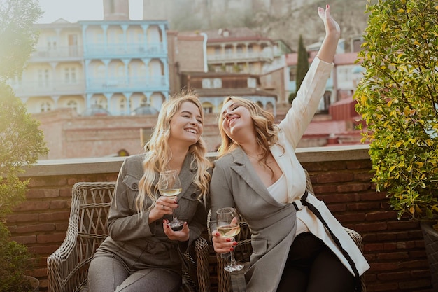 Closeup portrait of two female friends in strict suits laughing drinking wine on the terrace outside