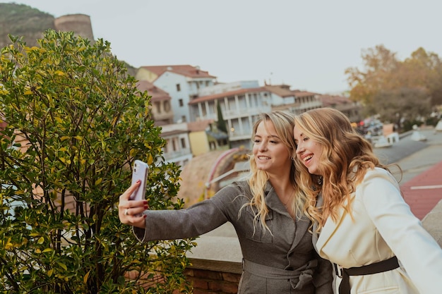 Closeup portrait of two female friends in strict suits laughing taking selfie on the terrace outside