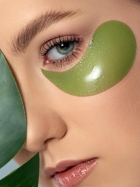 Closeup portrait of young beautiful woman with green eye patches under her eyes Girl hiding behind a leaf Skin treatment and morning routine concept