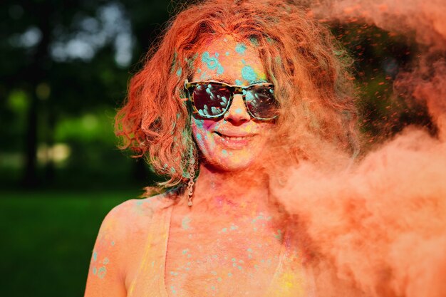 Closeup portrait of young caucasian woman with curly hair posing in a cloud of orange Gulal paint. Space for text