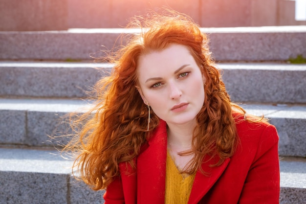 Closeup portrait young caucasian woman with red curly hair in red coat in wind