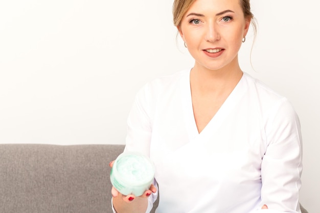 Closeup portrait of young smiling female caucasian healthcare worker standing with jar of cream beauty product on white background