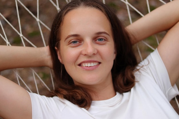 Closeup portrait of young woman with brown hair wearing casual white tshirt lying in hammock outdoors raised her arms behind head looking at camera relaxing in open air