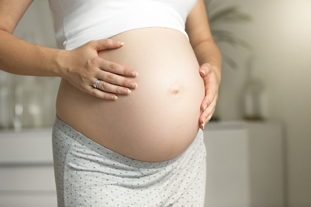 Closeup pregnant woman posing at window and holding hands on belly