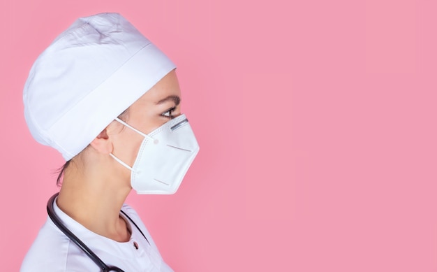 Closeup profile portrait of a young female doctor in a medical protective mask on a pink wall with a space for text