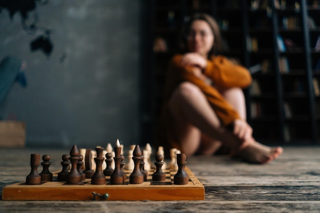 Closeup selective focus to chess board on wooden vintage floor blurred unrecognizable woman sitting in far near bookshelves in dark library room