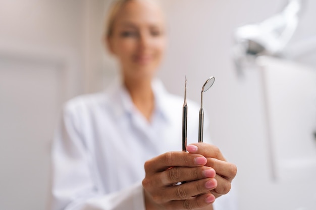 Closeup selective focus shot of female dental doctor holding working tools looking at camera in dentistry clinic with modern white interior Concept of stomatology dentistry and orthodontics