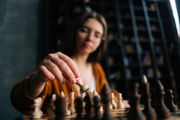 Closeup selective focus of smart young woman in elegant eyeglasses making chess move sitting in armchair in dark library room