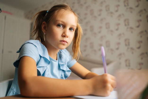 Closeup of serious primary little child girl writing doing homework sitting at home table by window