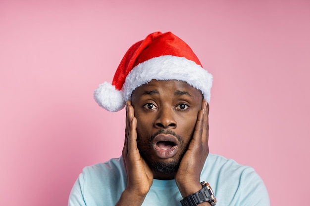 Closeup shot of attractive dark skinned man wearing Santa hat looks with surprisement, sees something stunning, keeps palms on cheeks, standing against pink wall. Facial expressions, reactions.