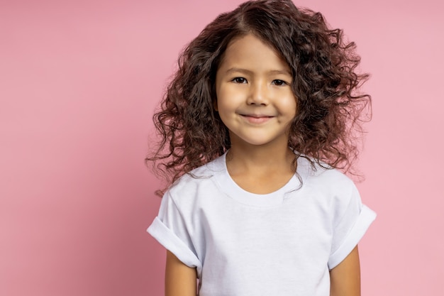 Closeup shot of charming caucasian little girl in white t shirt, with friendly, kind look looking with cute smile, posing against pink wall. Happy childhood, childish innocence, kids concept