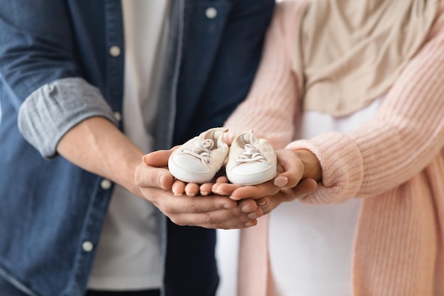 Closeup shot of expectant muslim family holding small baby shoes in their hands, unrecognizable pregnant islamic lady in hijab and her loving husband enjoying future parenthood, cropped image