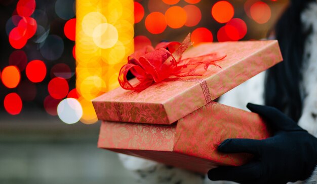 Closeup shot of female hands in gloves holding gift box at the background of red and yellow lights. Space for text