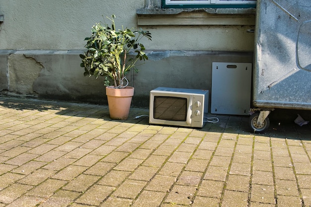 Closeup shot of an old microwave and a plant in a bucket on the ground next to the garbage box