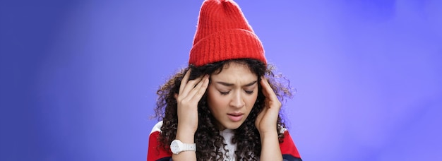 Closeup shot of sad and sick cute stylish woman in red warm beanie close eyes rubbing temples feelin