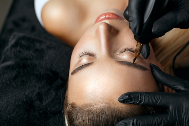 Closeup shot of a woman in black gloves applying permanent brow makeup to a young woman. Space for text