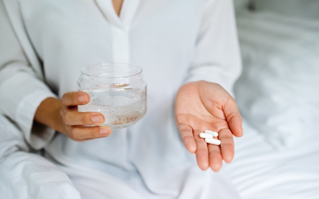 Closeup  of a sick woman holding white pills and a glass of water while sitting on a bed