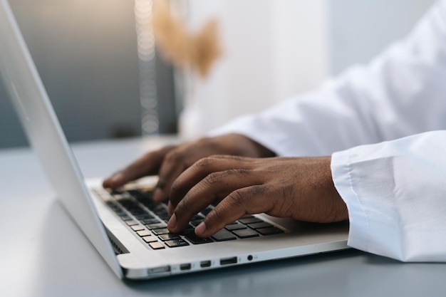 Closeup side view hands of unrecognizable AfricanAmerican male doctor wearing white coat working typing on laptop computer