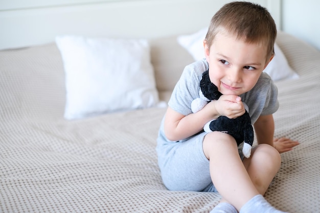 Closeup of a small happy caucasian boy in a gray tshirt lies on the bed with a toy dog