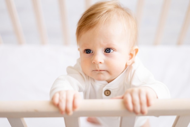 Closeup of a smiling baby 6 months old blond boy in a crib in a bright bedroom in a white cotton bodysuit portrait concept of children's goods