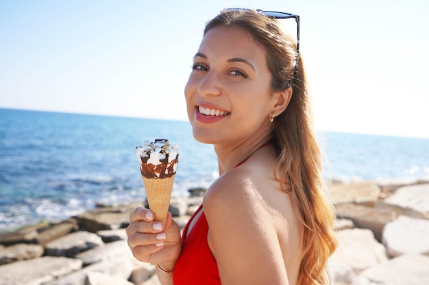 Closeup of smiling bikini woman holding ice cream cone italian gelato turns around to the camera on the beach on summer