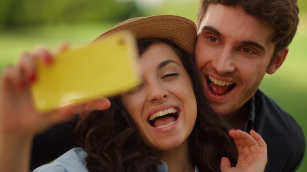 Closeup smiling couple having video call on mobile phone at picnic Portrait of handsome man biting woman ear outdoors Happy people saying hi on smartphone camera in summer park