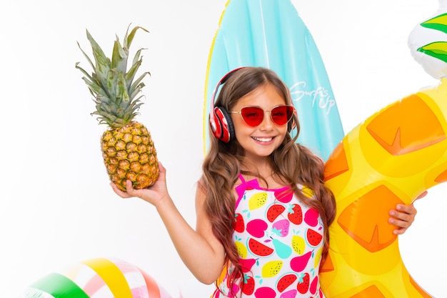 Closeup smiling girl on a white surface, the child holds a pineapple in his hands surrounded by swimming accessories, red headphones on his head