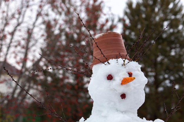 Closeup of snow man head with cones and carrot instead of eyes and nose flower pot on head and tree branches instead of hands and hair Having fun outside in winter